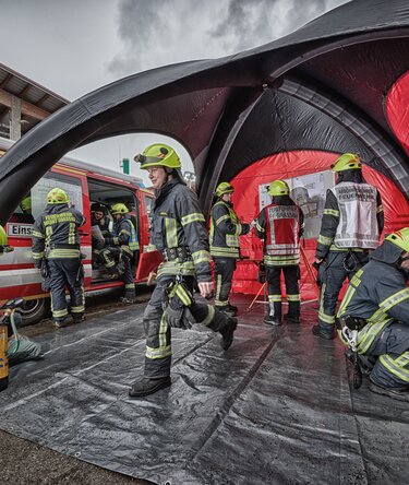 Firefighters working inside and in front of a red and black inflatable emergency tent next to an emergency vehicle.